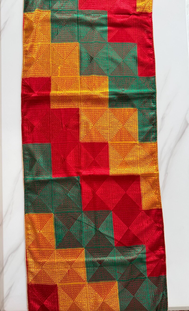 May include: A rectangular table runner featuring a geometric pattern of squares in vibrant red, emerald green, and golden yellow. The fabric displays a textured, stitched grid design across the entire surface, creating a repeating block motif.