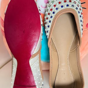 May include: Pair of embellished white flats with colorful gems and white beading. The soles are red, and the inner sole is tan. The shoes are displayed on a white surface with a plush orange pillow in the background.