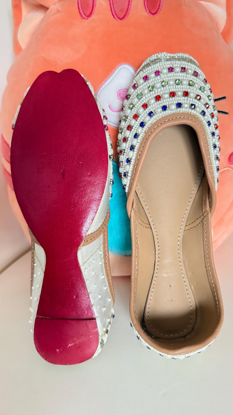 May include: Pair of embellished white flats with colorful gems and white beading. The soles are red, and the inner sole is tan. The shoes are displayed on a white surface with a plush orange pillow in the background.