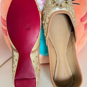 May include: A pair of beige and gold embellished slip-on shoes. The shoes have a red sole and are decorated with gold beads and embroidery. The interior is a light tan color. The shoes are likely for special occasions.