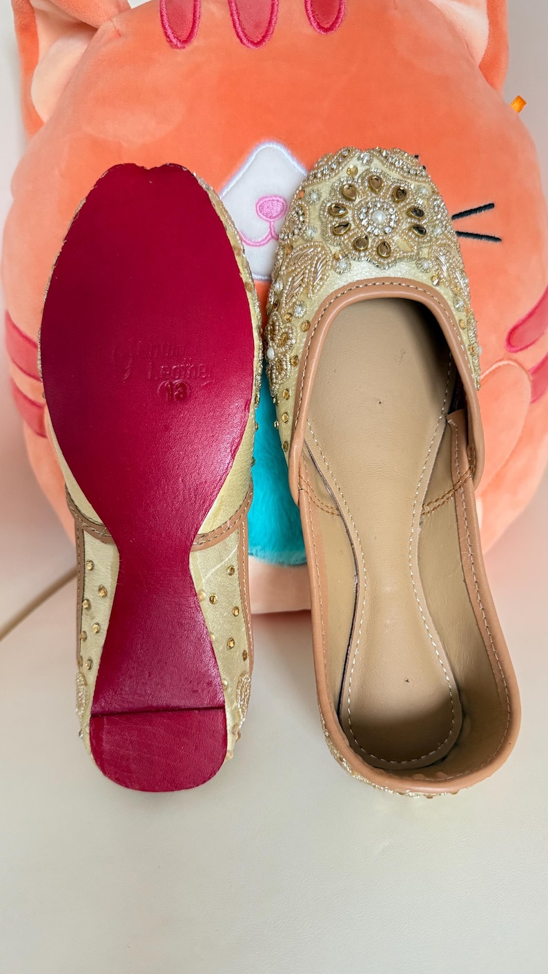 May include: A pair of beige and gold embellished slip-on shoes. The shoes have a red sole and are decorated with gold beads and embroidery. The interior is a light tan color. The shoes are likely for special occasions.