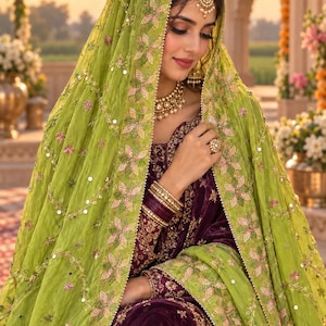 Op de afbeelding: Een dieppaars fluwelen kledingstuk met ingewikkeld goudborduurwerk, gecombineerd met een transparante limoengroene dupatta versierd met bloemmotieven, lovertjes en een gouden rand. Het ensemble bevat een gouden ketting, oorbellen en armbanden.