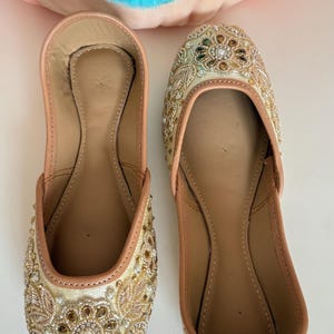 May include: A pair of beige and gold embellished slip-on shoes. The shoes feature intricate beadwork and embroidery in gold, brown, and clear tones. The interior is a light tan color, and the shoes are set against a white background.