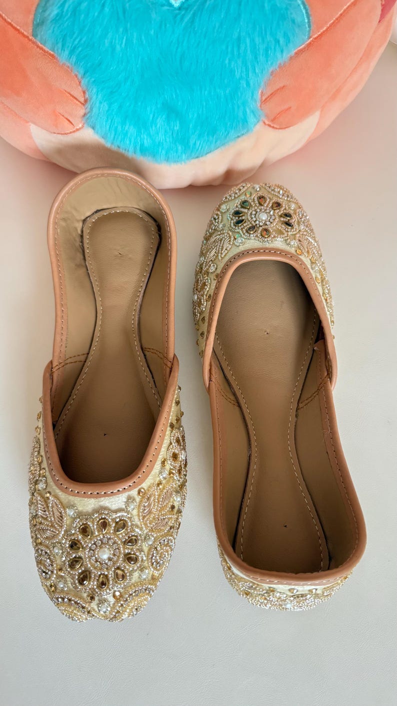 May include: A pair of beige and gold embellished slip-on shoes. The shoes feature intricate beadwork and embroidery in gold, brown, and clear tones. The interior is a light tan color, and the shoes are set against a white background.