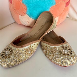 May include: A pair of cream-colored, embellished flats with intricate gold and pearl beadwork. The shoes have a pointed toe and a tan trim. A plush orange cat-shaped pillow with a blue heart is in the background.
