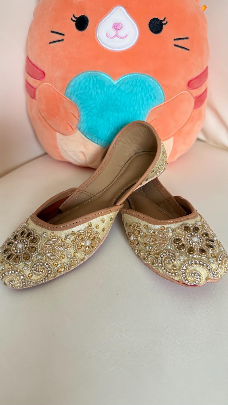 May include: A pair of cream-colored, embellished flats with intricate gold and pearl beadwork. The shoes have a pointed toe and a tan trim. A plush orange cat-shaped pillow with a blue heart is in the background.