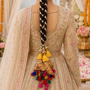 May include: A beige floor-length dress with intricate gold embroidery and pink floral accents, featuring long sleeves and a scoop back. A long black braided hairstyle is adorned with gold ribbon and a large yellow tassel accessory with colorful pom-poms.