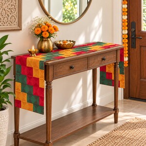 May include: A wooden console table with two drawers and a lower shelf, draped with a rectangular fabric runner featuring a geometric pattern of red, yellow, and green squares. A brass vase with orange flowers and a small brass oil lamp sit on top.