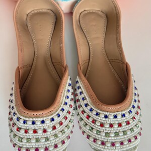 May include: A pair of embellished slip-on shoes. The shoes have a tan leather interior and a white beaded exterior, accented with red, blue, and green beads. The shoes are designed with a rounded toe and a flat sole.