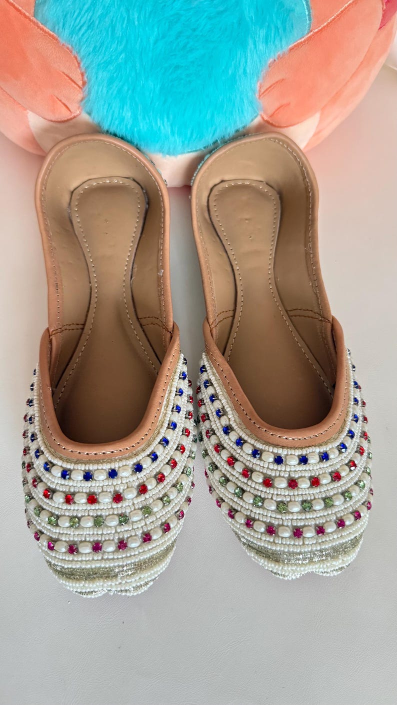 May include: A pair of embellished slip-on shoes. The shoes have a tan leather interior and a white beaded exterior, accented with red, blue, and green beads. The shoes are designed with a rounded toe and a flat sole.