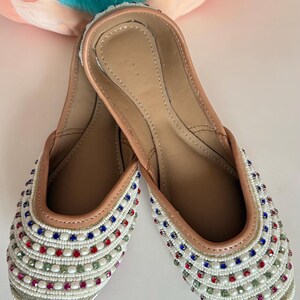 May include: White footwear with colorful bead embellishments. The shoes have a light brown interior and a white exterior adorned with red, blue, and green beads. The text on the image reads: Design 2 - White color footwear with colored beads.