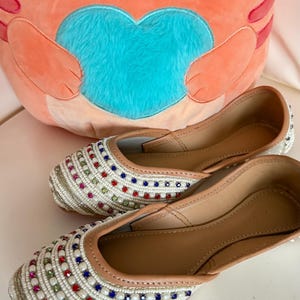 May include: A pair of white beaded flats with colorful square accents. The shoes have a tan leather interior and a cream-colored beaded exterior. A plush orange cat-shaped pillow with a blue heart is in the background.