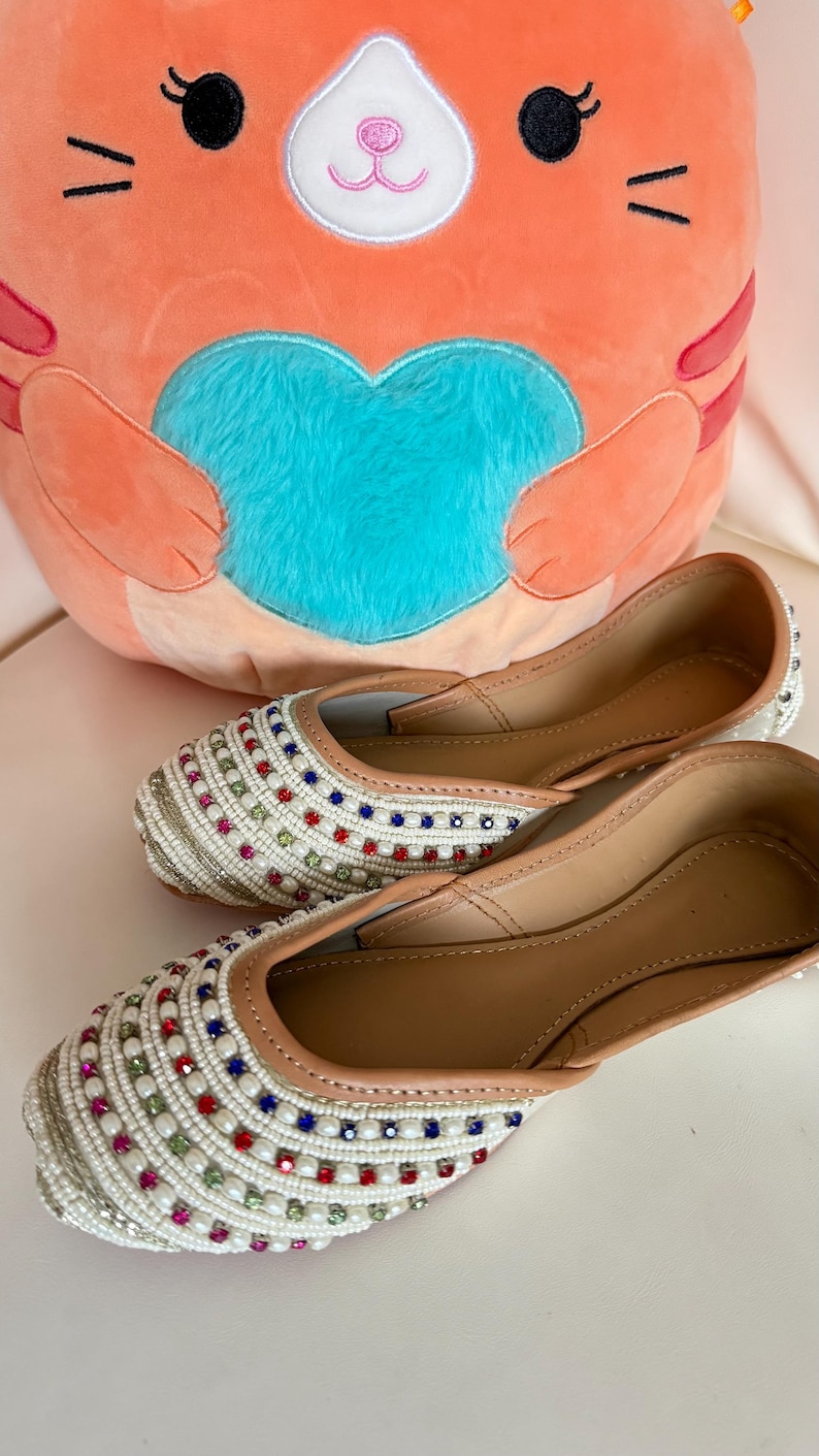 May include: A pair of white beaded flats with colorful square accents. The shoes have a tan leather interior and a cream-colored beaded exterior. A plush orange cat-shaped pillow with a blue heart is in the background.