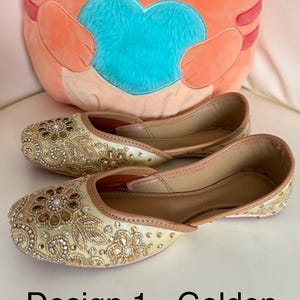 May include: A pair of gold-colored embroidered flats with intricate floral and jewel detailing. The shoes are displayed on a white surface, with a plush orange cat-shaped pillow in the background. Text on the image reads "Design 1 - Golden embroidery."