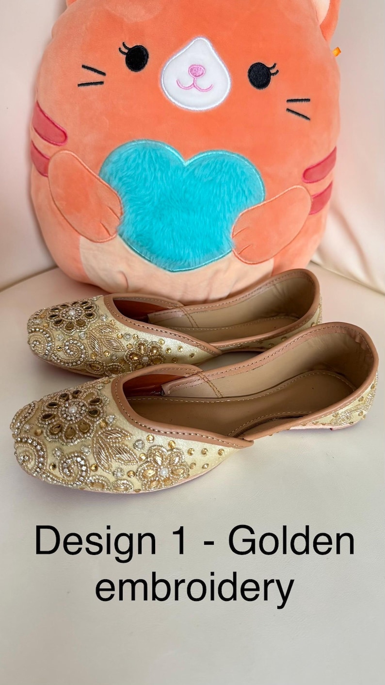 May include: A pair of gold-colored embroidered flats with intricate floral and jewel detailing. The shoes are displayed on a white surface, with a plush orange cat-shaped pillow in the background. Text on the image reads "Design 1 - Golden embroidery."