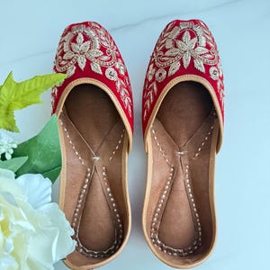 May include: A pair of red velvet flats featuring intricate gold thread embroidery on the rounded toe area. The shoes have a tan leather-colored interior lining and trim with visible stitching along the edges of the footbed and the outer sole.