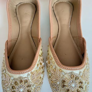 May include: A pair of cream-colored, embellished flats. The shoes feature intricate gold and brown beadwork and embroidery on the toe and sides. The inner sole is a light tan color, and the trim is a soft pink.