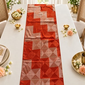 May include: A rectangular table runner featuring a vibrant orange fabric base with a repeating geometric pattern of metallic silver squares and triangles. The textile has a subtle sheen and is laid flat, showcasing a structured, symmetrical design.