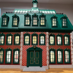 Op de afbeelding: Een adventskalender in de vorm van een bakstenen gebouw met een groen dak en groene afwerking. De structuur heeft vierentwintig genummerde ramen en deuren die als vakjes dienen. De gevel bestaat uit rode bakstenen muren, steenkleurige hoekaccenten en een centrale groene dubbele deur met het nummer vierentwintig erboven.