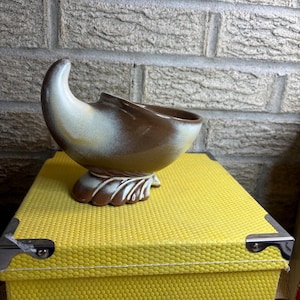 May include: A ceramic planter in a brown, cream, and yellow color scheme. The planter has a curved, horn-like shape with a decorative base. The planter is sitting on a yellow box with metal corner protectors. The background is a gray brick wall.