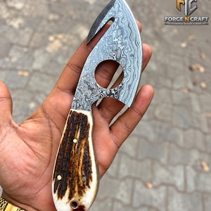 May include: A gut hook knife featuring a patterned Damascus steel blade with a circular cutout and a textured brown antler handle secured with three brass pins. The blade is etched with the text Forge N Craft.