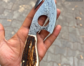 May include: A gut hook knife featuring a patterned Damascus steel blade with a circular cutout and a textured brown antler handle secured with three brass pins. The blade is etched with the text Forge N Craft.