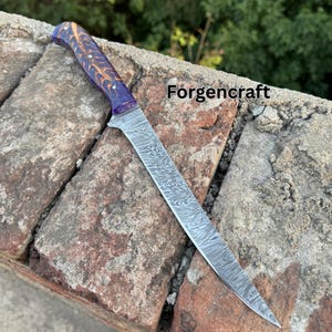 Puede incluir: Un cuchillo largo de acero de Damasco con una hoja estampada y un mango morado y dorado. El cuchillo está sobre una superficie de ladrillo, con el texto "Forgencraft" visible.