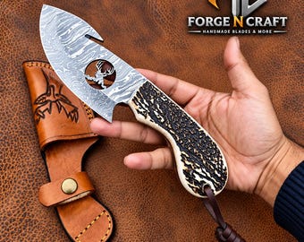 May include: A Damascus steel gut hook knife with a textured antler handle and a leather sheath. The blade features a circular cutout with a deer silhouette. A brown leather lanyard is attached to the handle. The logo Forge N Craft is visible.