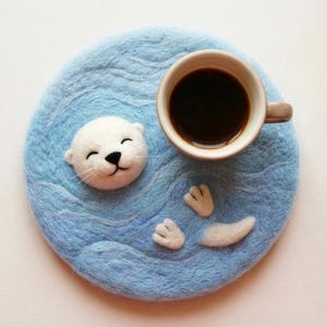 May include: A handmade blue felt coaster with a white seal design. The seal has a smiling face and is surrounded by a wavy pattern. A cup of coffee sits on the coaster, adding a functional and decorative touch.