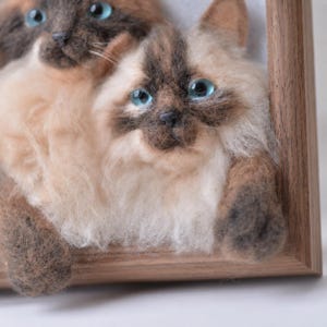 May include: A needle felted wall art piece featuring two Siamese-style cats with cream and dark brown fur, bright blue eyes, and textured wool details, set within a light brown wooden frame that highlights the three-dimensional cat faces.