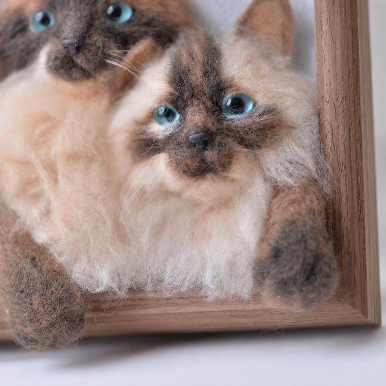 May include: A needle felted wall art piece featuring two Siamese-style cats with cream and dark brown fur, bright blue eyes, and textured wool details, set within a light brown wooden frame that highlights the three-dimensional cat faces.