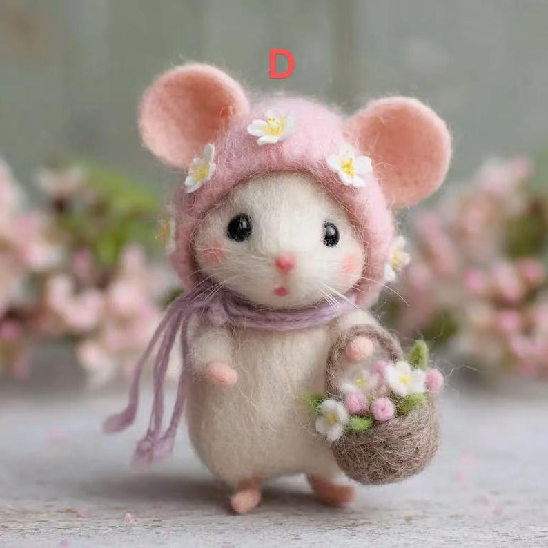May include: A handcrafted felt mouse figurine wearing a pink bonnet adorned with white flowers and a matching scarf. The mouse holds a small woven basket filled with flowers. The figurine has large black eyes and pink cheeks.