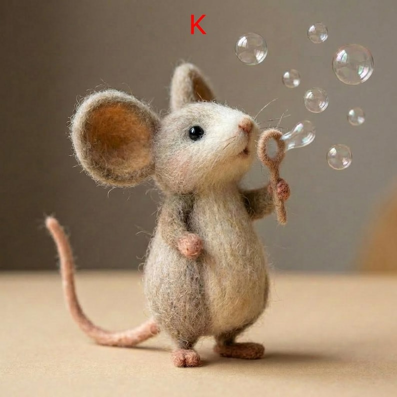 May include: A handcrafted felt mouse figurine blowing bubbles. The mouse is light brown with large ears and a long tail. It holds a bubble wand, and several clear bubbles float in the air. The letter "K" is in the upper left corner.