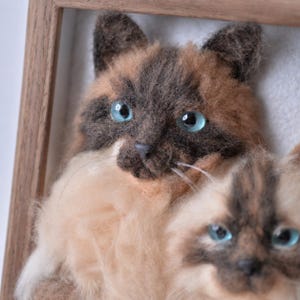 May include: A needle-felted wall art piece featuring two cats with light tan and dark brown fur, bright blue glass eyes, and white whiskers, encased in a simple wooden frame. The textured fiber art depicts the feline faces in detail.