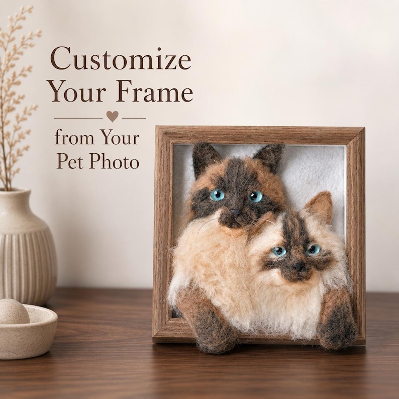 May include: A square wooden frame containing a needle-felted 3D portrait of two cats with blue eyes and brown and cream fur. The text Customize Your Frame from Your Pet Photo is printed in brown above the frame next to a small vase.