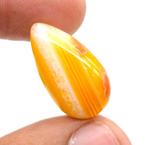 May include: A teardrop-shaped gemstone in shades of yellow and orange, with white and reddish-orange streaks. The polished stone is held between two fingers against a white background, highlighting its natural patterns and translucence.