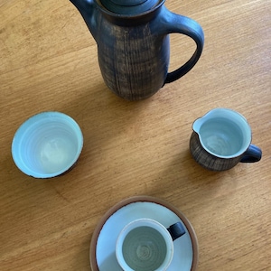 May include: A ceramic tea set with a dark brown and black textured teapot, a small creamer, a teacup on a saucer, and a small bowl. The interior of the teacup, creamer, and bowl are light blue.