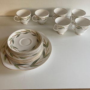 Op de afbeelding: Een vintage theeservies met zes witte kopjes met oren, gouden rand en een groen bladerenpatroon. De set bevat een stapel schotels en een serveerschaal. De kopjes en schotels zijn gerangschikt op een wit oppervlak.
