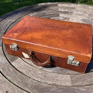 May include: A vintage brown leather suitcase with metal latches and a handle. The suitcase is rectangular and shows signs of wear, with a slightly distressed appearance. It sits on a wooden table in natural sunlight.