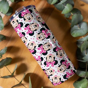 K&ouml;nnte beinhalten: Ein Edelstahlbecher mit einem sich wiederholenden Muster aus Minnie Mouse-Gesichtern, rosa Schleifen mit Tupfen und schwarzen Akzenten auf wei&szlig;em Hintergrund. Der Becher ist zylindrisch und hat einen transparenten Deckel.