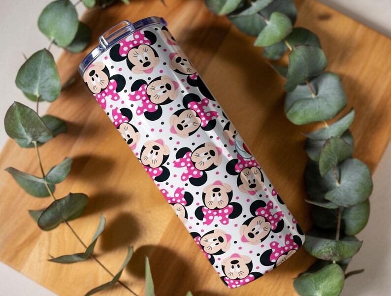 K&ouml;nnte beinhalten: Ein Edelstahlbecher mit einem sich wiederholenden Muster aus Minnie Mouse-Gesichtern, rosa Schleifen mit Tupfen und schwarzen Akzenten auf wei&szlig;em Hintergrund. Der Becher ist zylindrisch und hat einen transparenten Deckel.