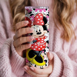 K&ouml;nnte beinhalten: Ein Edelstahlbecher mit einem farbenfrohen Minnie Mouse-Design. Der Becher zeigt Minnie in ihrem klassischen roten Polka-Dot-Kleid und ihrer Schleife. Der Hintergrund ist wei&szlig; mit rosa Punkten und Minnie Mouse-Silhouetten.