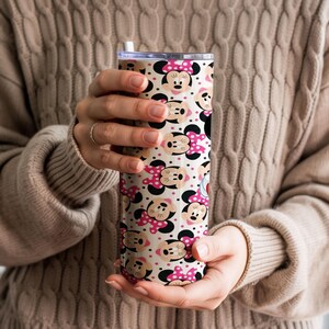 K&ouml;nnte beinhalten: Ein Edelstahlbecher mit einem sich wiederholenden Muster von Minnie Mouse-Gesichtern mit rosa Schleifen auf cremefarbenem Hintergrund. Der Becher hat einen transparenten Deckel und einen wei&szlig;en Strohhalm. Das Design ist verspielt und f&uuml;r Getr&auml;nke geeignet.