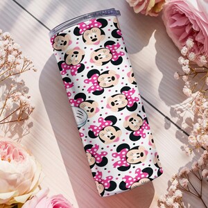 K&ouml;nnte beinhalten: Ein wei&szlig;er Becher mit einem sich wiederholenden Muster aus Minnie-Mouse-Gesichtern, rosa Schleifen und schwarzen Punkten. Der Becher hat einen transparenten Deckel. Rosa Rosen und wei&szlig;e Blumen umgeben den Becher.