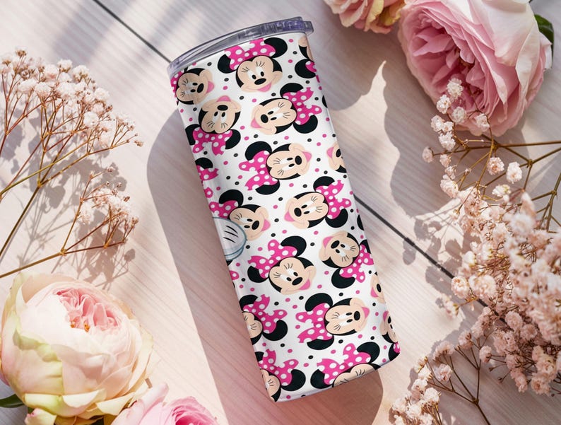 K&ouml;nnte beinhalten: Ein wei&szlig;er Becher mit einem sich wiederholenden Muster aus Minnie-Mouse-Gesichtern, rosa Schleifen und schwarzen Punkten. Der Becher hat einen transparenten Deckel. Rosa Rosen und wei&szlig;e Blumen umgeben den Becher.