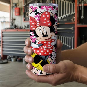 K&ouml;nnte beinhalten: Ein Edelstahlbecher mit Minnie Maus. Der Becher hat einen wei&szlig;en Hintergrund mit einem Muster aus Minnie Maus-Gesichtern und rosa Tupfen. Minnie Maus tr&auml;gt ein rotes Kleid mit wei&szlig;en Punkten und eine passende Schleife.