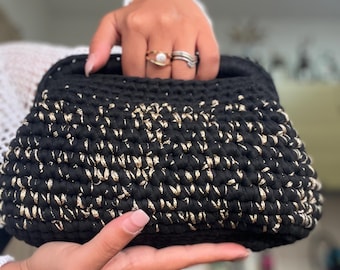 May include: A black and gold crochet handbag with a handle. The bag is held by a person. The crochet pattern features a mix of black and gold threads, creating a textured appearance. The bag's shape is rectangular with rounded edges, suitable for carrying essentials.