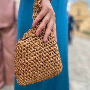 Può includere: Una borsa intrecciata, marrone dorato, con un manico abbinato. La borsa ha un aspetto strutturato, simile a un cestino, ed è tenuta contro un vestito color verde acqua. Il manico è avvolto attorno al polso. La borsa ha una forma rettangolare arrotondata.
