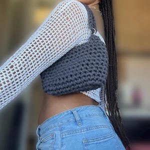 Può includere: Una borsa a tracolla all'uncinetto grigio scuro con un anello in metallo color argento sulla tracolla. La borsa è tenuta contro una maglia bianca a maniche lunghe a trama aperta e jeans blu chiaro.