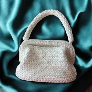Può includere: Una borsa bianca intrecciata con un manico curvo. La borsa ha una forma rettangolare con una superficie strutturata. Il manico è anch'esso intrecciato e arcuato. La borsa è appoggiata su un tessuto color verde acqua, simile al raso.